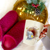 Person holding a mug with a bear design and text, surrounded by pink mittens and a decorative wreath.