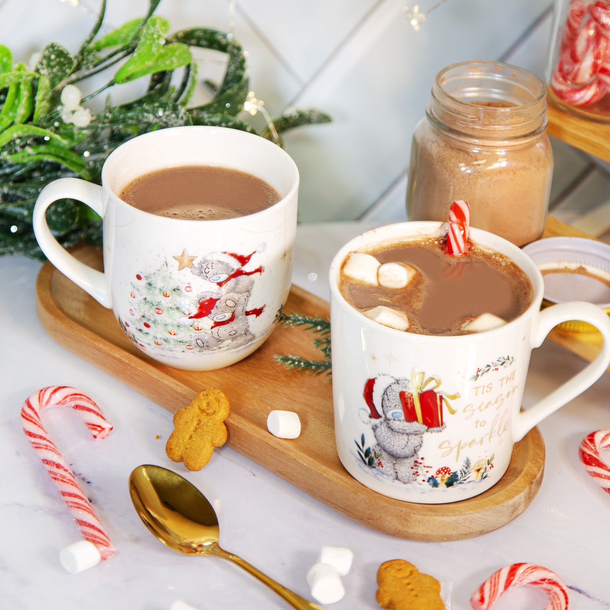 Two white ceramic mugs filled with hot cocoa on a wooden tray, surrounded by candy canes, marshmallows, gingerbread cookies, and a gold spoon in a cozy holiday setting.