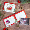 A styled flat-lay of a Tiny Tatty Teddy My 1st Christmas Memories Book  and on a rustic wooden table, surrounded by holiday greenery, red berries, scissors, and washi tape. The book cover shows Tiny Tatty Teddy in a sleigh, and the open interior page has a photo placeholder area.